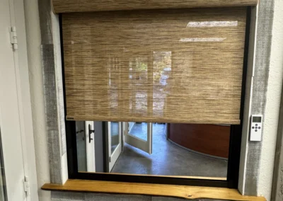 A window with bamboo blinds partially drawn, framed by wood and textured gray wall, showing a glimpse of an interior space.