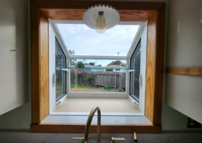 View through a kitchen window, showcasing a garden, wooden fence, and cloudy sky, framed by wooden trim and a pendant light above.