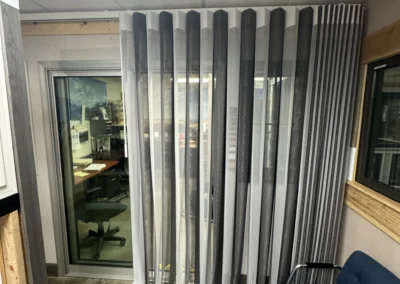 A corner office features vertical sheer and opaque blinds, with a blue chair beside a glass door leading to an office space.